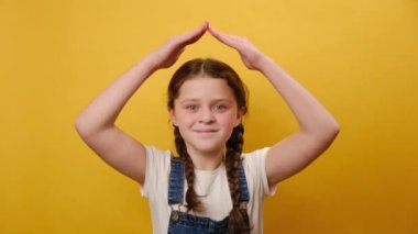Sevimli gülümseyen reşit olmayan kız çocuğunun portresi ellerini çatı gibi başının üstünde tutarak, evde kalıp stüdyodaki sarı arka plan duvarının üzerinde izole bir şekilde poz veriyor. Çocukluk yaşam tarzı konsepti