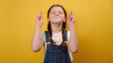 Sevimli çocuk portresi özel anı bekliyor, parmaklarını çapraz yapıyor, dilek tutuyor, gözleri kapalı, stüdyodaki sarı arka plan duvarının üzerinde izole edilmiş. Çocukluk yaşam tarzı konsepti