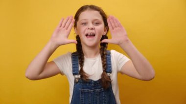Stüdyodaki sade sarı arkaplan duvarının üzerinde izole bir şekilde poz veren, ağza yakın elleri olan, eğlenceli, gizemli, küçük kız çocuk çığlık haberlerinin portresi. Çocukluk yaşam tarzı konsepti