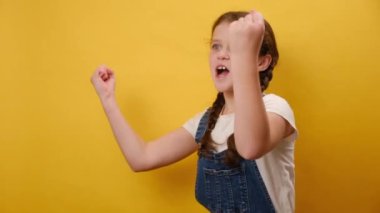 Heyecanlı küçük kız çocuğunun portresi, stüdyodaki sade sarı arkaplan duvarının üzerinde izole edilmiş pozlar vererek kazanan yumruk hareketi yapıyor. Çocukluk yaşam tarzı konsepti