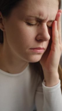 Oturma odasındaki kanepede oturan yorgun, sağlıksız genç bir kadının dikey videosu. Gözleri kapalı, el ele tutuşmuş, şiddetli baş ağrısı ya da migrenden muzdarip, zonklayan ağrı konsepti.