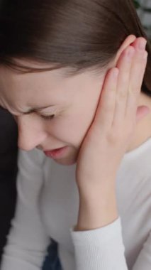 Üzgün, genç, esmer bir kadının acı dolu kulağını tutarken aniden güçlü bir ağrı hissettiği dikey bir video. Sağlıksız beyaz kadın evdeki kanepede oturan acı veren otit iltihabından muzdarip. Sağlık sorunları kavramı