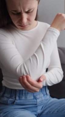 Dirsek ağrısı olan, evde koltukta masaj yapan, sağlıksız, üzgün, beyaz bir kadının dikey görüntüleri. Sağlık, eklem iltihabı dirsek kavramında ağrı