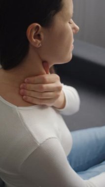 Yorgun, beyaz, boynu ağrıyan, boynu ağrıyan, vücudu ağrıyan, masaj yapan, omzu ağrıyan, evdeki koltukta oturan genç bir kadının dikey görüntüleri. Ağrı vücut kasları sert sorun