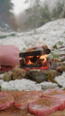 Erkek elinin çiğ bifteğe tuz serptiği ve pişirmeye hazırlandığı dikey görüntüler. Kış zamanı kamp ateşinin yanında marine edilmiş et. Kamp ortamı ve açık hava yaşam tarzı konsepti