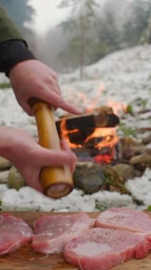 Erkek eli, kış ormanlarında şenlik ateşinin yanında çiğ biftek üzerine baharat serpiyor. Kamp ateşinde yemek pişirmek. Kamp ortamı ve açık hava yaşam tarzı konsepti