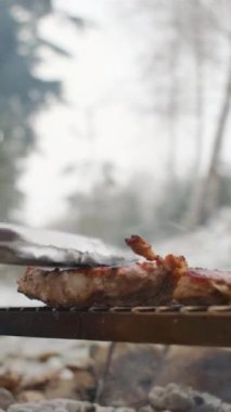 Genç adamın kış vakti ormanda biftek ve metal ızgara pişirirken çekilmiş dikey videosu. Kamp ateşinde yemek pişirmek. Kamp ortamı ve açık hava yaşam tarzı konsepti