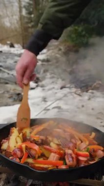 Adamın iştah açıcı patatesleri tavada kızartılmış sebzelerle pişirdiği dikey video dağ kömürü, nehir ve orman yakınlarında. Erkek eli kışın yemek karıştırıyor.