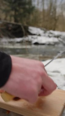 Arka plandaki kış nehrinin tahta tahtasında erkeklerin sosis doğrarken çekilmiş dikey görüntüleri. Lezzetli organik domuz eti dilimleyen adam köy kış pikniği yapıyor. Açık hava yemeklerini düşün.