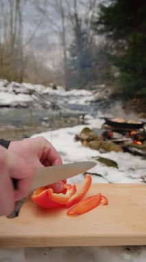 Kışın şenlik ateşi ve dağ nehri yakınlarındaki tahta tahtada taze kırmızı biber kesen adamın dikey videosu. Yemek pişirme, macera aktif tatiller, kamp ortamı, açık hava yaşam tarzı.
