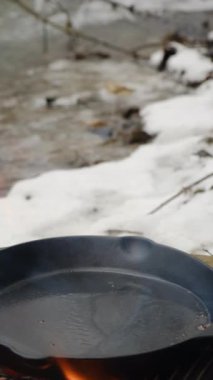 Bilinmeyen bir gencin kış zamanı dağların kenarındaki nehirde kızartma tavasında patates pişirdiği dikey görüntüler. Yürüyüşte dışarıda yemek pişirmek. Yiyecek konsepti