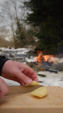 Erkek ellerinin taze patates bıçağını kestiği dikey video arka plandaki şenlik ateşinde ve kış zamanı dağlardaki nehirde. Yemek pişirme konsepti