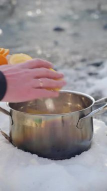 Erkek elinin dikey görüntülerine göre elmalar, portakallar kışın açık havada sıcak şarap pişirmek için. Geleneksel içeceklerin ısınma süreci, karla kaplı nehir kenarındaki şenlikli atmosfer.