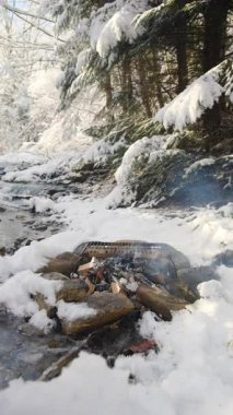Genç beyaz adamın şenlik ateşine ot koyduğu, dağların kenarında şarap pişirdiği ve kış vakti karla kaplı harika bir ormanın olduğu dikey bir video. Kamp ateşinde sıcak içecekler pişiriyorum.