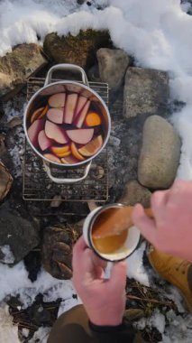 Genç adamın dikey görüntülerine bal ekleniyor portakallı, elmalı, karanfilli, anasonlu ve tarçınlı sıcak şarabın kış mevsiminde kamp ateşinde pişirildiği şarap. Açık havada renkli şenlikli sıcak içecekler