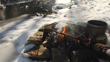 Erkek eli, kış vakti arka plandaki nehirde kamp ateşinin üzerine su dolu kazan koyuyor. Kış mevsiminde şenlik ateşi. Macera, seyahat ve yürüyüş kavramı. Hızlı yemek kampı.