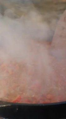 Doğada şenlik ateşinde tavada çürüyen kırmızı biberli domates ve baharat pişiren genç adamın dikey videosu. Kamp ortamı, doğa ve açık hava yaşam tarzı. Yemek pişirme konsepti