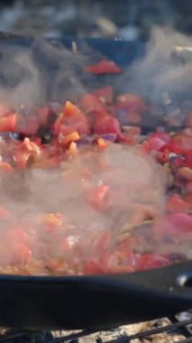 Adamın kışın açık havada tavada sebze kızarttığı dikey video. Sağlıklı yemek, diyet, düşük kalori. Doğranmış domatesler soğan ve kırmızı biberle kızartma tavasına düşer. Şenlik ateşinde sebze sote