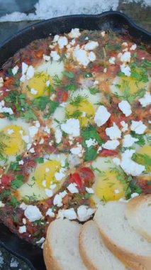 Ekmek ve Shakshuka 'nın kış zamanı arka plandaki vahşi nehir üzerindeki dikey görüntüleri. Shakshuka yumurtaları, domates, büyük taşta sıcak tavada biber. Kamp ortamı, açık hava yaşam tarzı konsepti.