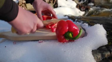 Genç adamın kışın, arka plandaki dağlar nehrinin tahta tahtasında taze kırmızı biber keserken yakın plan görüntüsü. Yemek pişirme, macera aktif tatiller, kamp ortamı, açık hava yaşam tarzı.