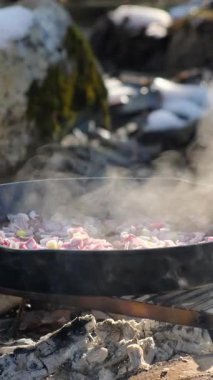 Genç adamın kışın açık havada tavada sebze kızarttığı dikey video. Sağlıklı yemek, diyet, düşük kalori. Biber dilimleri tavaya soğanla düşer. Şenlik ateşinde sebze sote