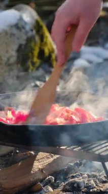 Bilinmeyen bir gencin dikey videosu, kış zamanı dağlar nehrinin yanındaki şenlik ateşinde sebzeleri karıştırıyor. Yemek pişirme konsepti