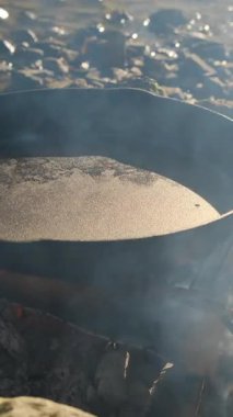 Bilinmeyen bir gencin dikey videosu, ilkbaharın başlarında nehrin kenarında tavaya zeytinyağı döküyor. Dinlen ve doğada piknik yap. Yemek pişirme konsepti. Kamp yaşam tarzı rutini