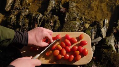 Sebze dilimlemek ve dışarıda yemek pişirmek. İnsan görünümünün en üst görüntüsü, ilkbaharın başlarında nehrin yanındaki tahta tahtada domatesleri keser. Taze salata vejetaryen mutfağı pişirmek için sebze dilimliyorum.