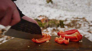 Kışın şenlik ateşi ve dağ ormanlarının yakınındaki tahta tahtada taze kırmızı biber kesen adama yaklaş. Yemek pişirme, macera aktif tatiller, kamp ortamı, açık hava yaşam tarzı.