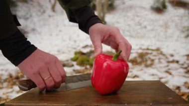 Kış zamanı kamp ateşi ve dağ ormanlarının yakınındaki tahta tahtada taze kırmızı biber kesen adama yaklaş. Yemek pişirme, macera aktif tatiller, kamp ortamı, açık hava yaşam tarzı.