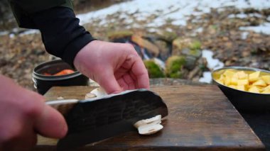 Meçhul genç adam kış veya ilkbahar başında kamp ateşinin yanında tahta tahtada bıçakla şampiyon mantarlarını kesiyor. Lezzetli yemekler için malzemeleri doğramak. Diyet sağlıklı yaşam tarzı konsepti