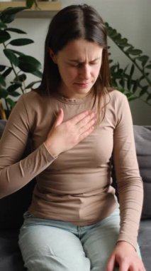 Kanepede tek başına otururken göğsü ağrıyan sağlıksız genç beyaz bir kadının dikey videosu. Üzgün bir kadın evinde kalp ağrısı, enfarktüs ya da kalp hastalığı konseptinden muzdarip.