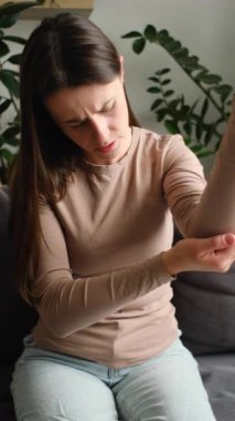 Dirsek ağrısı olan, evde koltukta yalnız başına masaj yapan, sağlıksız, üzgün, beyaz bir kadının dikey videosu. Sağlık, eklem iltihabı dirsek kavramında ağrı