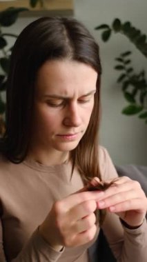 Kanepede tek başına oturan mutsuz, uzun saçlı genç bir kadının elinde kaybettiği saçlara bakarken çekilmiş dikey videosu. Saç dökülmesi, saç dökülmesi, kadın konsepti semptomları ve nedenleri