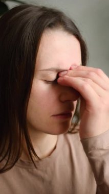 Yorgun, sağlıksız, genç bir kadının dikey görüntüsü. Kanepede tek başına oturuyor. Gözleri kapalı, el ele tutuşmuş, şiddetli baş ağrısı ya da migren ağrısı çekiyor.