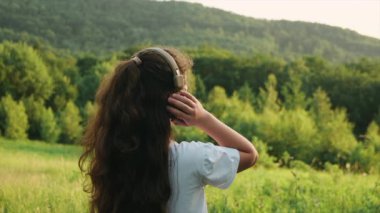 Neşeli genç kız kulaklıkla müzik dinliyor ve arka planda inanılmaz sıcak bir günbatımının tadını çıkarıyor inanılmaz dağlarda. Çocukluk ve yaz hafta sonu konsepti