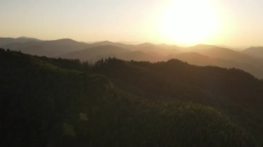 Güzel dağların havadan çekilmiş görüntüsü ve sıcak, inanılmaz gün batımı. Dağ sırasının yeşil orman yamaçları. Gökyüzündeki parlak güneş diski yazın yavaşça dağların tepesine çıkar. Doğa konsepti