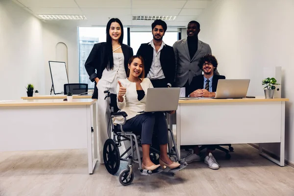 Office workers and woman on a wheelchair in bright office. They are showing a teamwork. Portrait of diverse business team with young woman in wheelchair all smiling at camera in office.