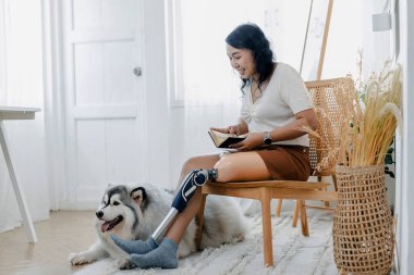 Protez bacaklı bir kadın evdeki köpeğin yanındaki sandalyede oturuyor. Evde köpeğiyle sandalyede oturan bir kadın. Oturma odasında oturan protez bacaklı çift..