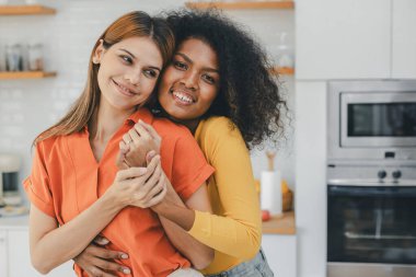 Mutfak odasında kucaklaşan çok ırklı lezbiyen çift. Yüzük tutan siyah beyaz bir çift. Afro-Amerikan ve beyaz lezbiyen çift birbirlerine şefkatle sarılıp bakıyorlar.