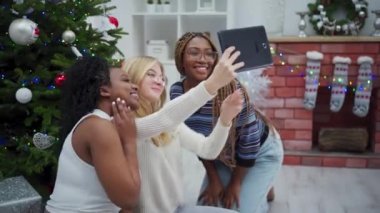 Kız arkadaşlar Noel günü buluşup Noel ağacının yanında bir tabletle selfie çekiyorlar. Gülümseyen ve mutlu kızlar.