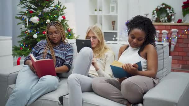 一个非洲女人在看书 一位欧洲妇女正在平板电脑上浏览网页 姑娘们在笑 圣诞节装饰的客厅 — 图库视频影像