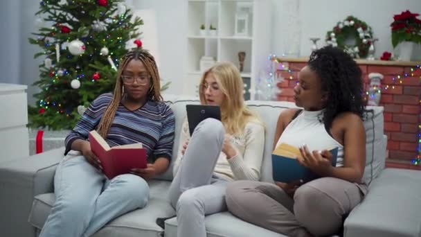 アフリカ人女性が本を読む ヨーロッパの女性がタブレットでウェブを閲覧しています 女の子たちは笑っている クリスマスのために装飾されたリビングルーム — ストック動画
