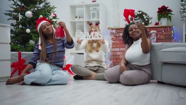 女の子は手にクリスマスのシンボルを保持します クリスマスの銘文 笑顔アフリカとヨーロッパの女性はリビングルームの床に座っています — ストック動画