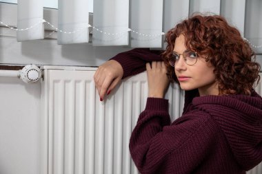 Radyatörün yanında oturan üzgün kadın. Hatalı merkezi ısıtma. Apartman bloklarında ısınma sezonu başlıyor..