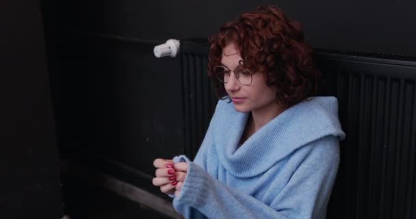 一个女孩坐在一个炽热的散热器旁取暖 年轻女子的肤色和卷曲的头发呈浅色 冬季中央供暖 — 图库视频影像