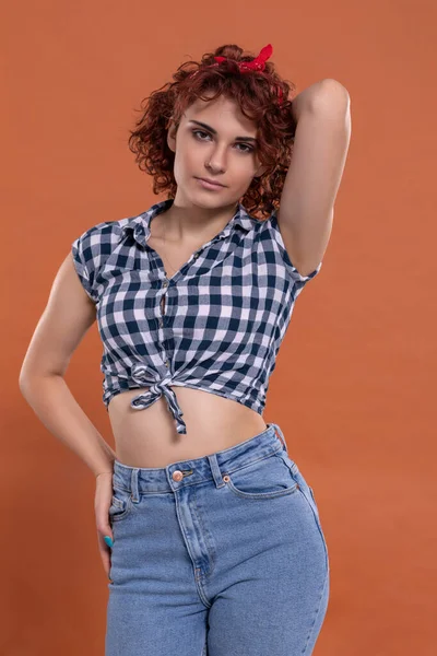 Young girl dressed in pin up style. Checkered shirt. Isolated from a solid background of brick color. Short curly hair in chestnut color.