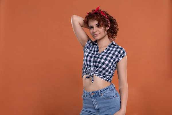 Young girl dressed in pin up style. Checkered shirt. Isolated from a solid background of brick color. Short curly hair in chestnut color.