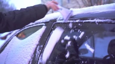 A woman clears snow from the side window of her car. The charms of the winter season when there is a lot of snow. A layer of wet snow on the car and car windows.