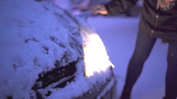 汽车前灯亮着 一个女人从客车前灯上擦去积雪 在冬季 女人穿着冬衣和围巾 — 图库视频影像